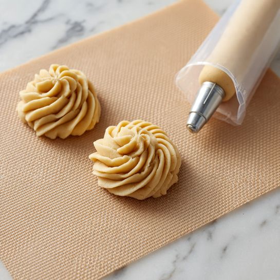 The image shows two swirled dollops of light beige dough or batter on a textured beige baking mat. Each dollop has a ridged, rosette shape with smooth, curved lines formed from a piping bag. To the right, there is a clear piping bag filled with the same light beige dough, held close to the mat and fitted with a metal star-shaped nozzle, slightly squeezing out some dough at the tip. The background surface is a white marbled texture. photo taken with an iphone --ar 4:5 --v 7