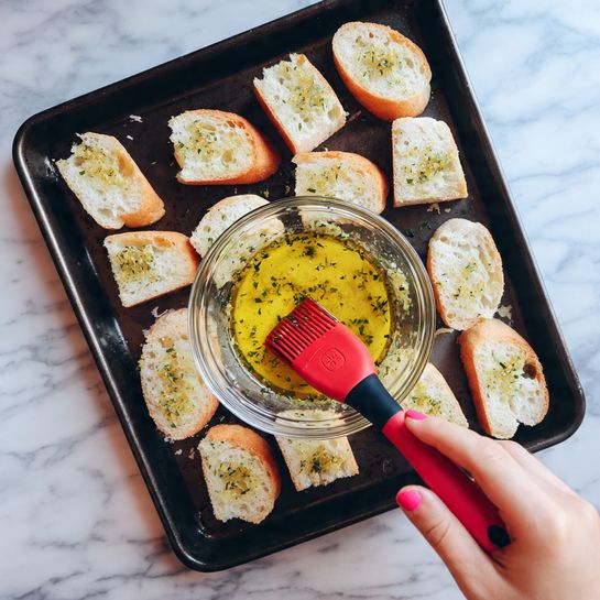 A woman's hand with pink nail polish is holding a clear glass bowl filled with a yellow oily mixture with green herbs, and a red silicone brush with a black handle is resting inside the bowl. Below the bowl, a dark baking tray holds sixteen small rectangular slices of white bread, arranged neatly in four rows and four columns, on a white marbled surface. The bread slices have a soft texture and golden brown crusts visible on some edges. photo taken with an iphone --ar 4:5 --v 7