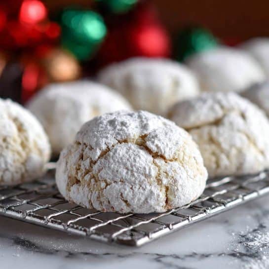 The image shows several small round cookies covered in white powdered sugar, arranged in rows on a silver wire cooling rack. The cookies have a rough, uneven surface texture, and the powdered sugar coating makes them look soft and snowy white. The rack is placed on a white marbled surface. The background is softly blurred with colors of red, green, and brown suggesting holiday decorations. The focus is sharp on the cookies in the front, highlighting their powdery texture. Photo taken with an iphone --ar 4:5 --v 7
