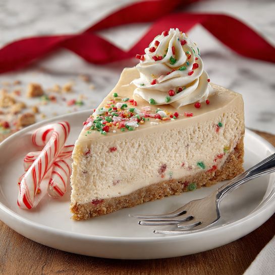 A slice of light tan cheesecake with a thick base made of crumbly, colorful bits sits on a white plate. On top of the cheesecake, there is a small swirl of white cream with red and green sprinkles, and more red and green sprinkles are scattered over the surface. Next to the cheesecake slice, there is a silver fork with a small piece of cheesecake on it. Two small candy canes with red and white stripes lay on the plate near the cheesecake. The background shows a wooden surface with a blurred red ribbon and crumbs, and the plate is placed on a white marbled texture. photo taken with an iphone --ar 4:5 --v 7