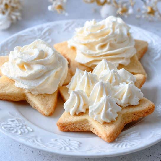 Three star-shaped cookies with light golden brown color are on a white plate with detailed raised patterns around the edge. Each cookie has a thick layer of white, creamy frosting on top, with two cookies showing smooth, swirled peaks and the one in front showing multiple small dollops closely placed together, giving it a fluffy texture. The background shows a white marbled surface. photo taken with an iphone --ar 4:5 --v 7
