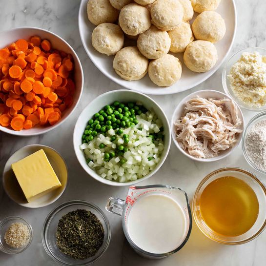 Creamy Chicken and Biscuits Soup with Fluffy Dumplings Recipe 4 This image shows several small white bowls arranged in a circle on a white marbled surface, each containing a different ingredient. One bowl has bright orange carrot slices, another has fresh green celery pieces, and a third holds diced white onions. There are frozen green peas filling one bowl, and shredded cooked chicken in another. A larger white plate holds round, pale biscuit dough pieces stacked in a circular pattern. Small clear glass bowls contain a yellow cube of butter, a mix of dried herbs and chopped garlic, a light tan flour, and one bowl with a golden broth. A glass measuring cup filled with white milk or cream is placed in front. All ingredients are organized neatly with bright natural light. Photo taken with an iphone --ar 4:5 --v 7