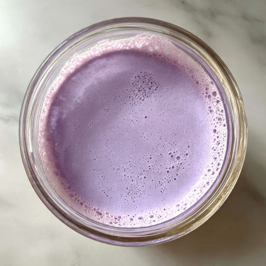 A close-up top view of a clear glass bowl filled with a smooth, light purple creamy mixture. The mixture has even texture with a few small bubbles and specks on the surface, and some splashes of the purple mixture stick around the rim of the bowl. The bowl is placed on a white marbled surface. photo taken with an iphone --ar 4:5 --v 7