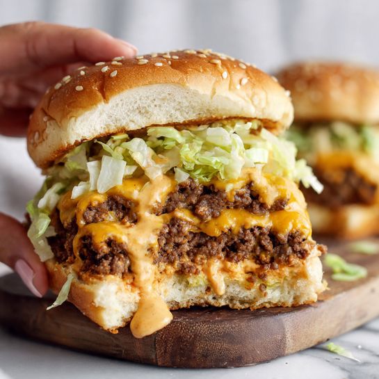 A close-up view of a burger held by a woman's hand showing three layers: the top soft sesame seed bun in golden brown, a middle layer of light green shredded lettuce mixed with melted pale orange cheese sauce, and a bottom thick layer of cooked ground beef that looks juicy with bits of cheese sauce dripping. The inside of the bun is soft and fluffy white, and the background shows more of the same burger on a wooden board on a white marbled surface. Photo taken with an iphone --ar 4:5 --v 7