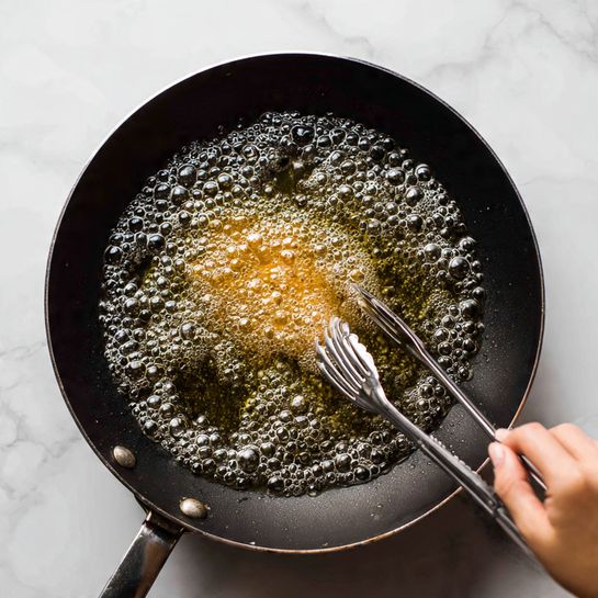 A dark cooking pan filled with bubbling clear oil showing active frying. In the middle, a light golden strip of food is frying, surrounded by many small bubbles. At the top right, a pair of metal tongs held by a woman's hand is about to hold the food. The pan sits on a black stove, with the setting against a white marbled texture. Photo taken with an iphone --ar 4:5 --v 7