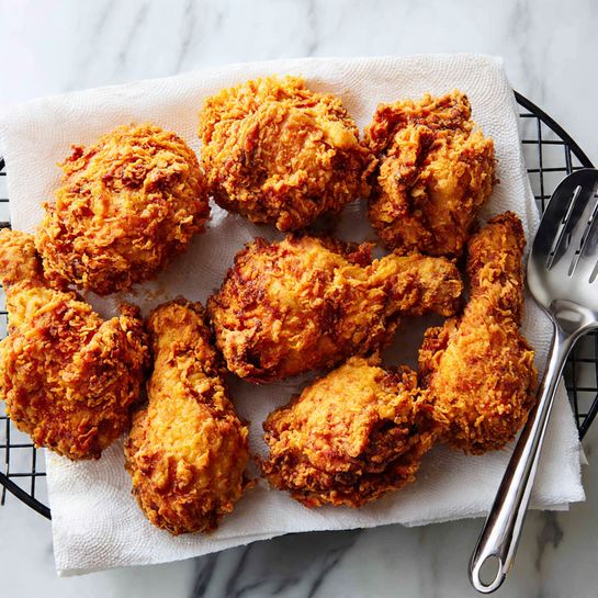 The image shows around fourteen pieces of golden brown fried chicken with a crispy, textured coating. The pieces vary in size and shape, some round and thick, others longer and thinner. The chicken sits on a white paper towel that lines a black metal cooling rack with a grid pattern. A silver spatula with a black handle is placed under some pieces on the right side. The background is a white marbled texture. The chicken has a crunchy look with some browned spots. Photo taken with an iphone --ar 4:5 --v 7