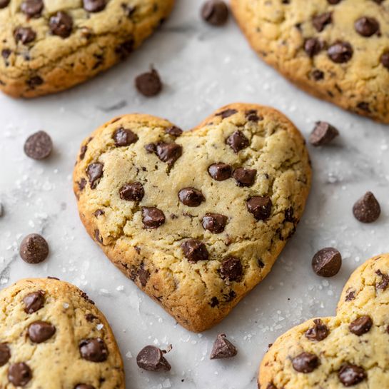 A white round plate holds seven heart-shaped cookies. Each cookie has a golden-brown edge with a light tan center, sprinkled with large, dark chocolate chips spread unevenly on top. The cookies have a soft, slightly bumpy texture with some melted chocolate chips adding a shiny contrast. The plate is placed on a white marbled surface, and the photo is taken from above, showing all cookies clearly. photo taken with an iphone --ar 4:5 --v 7