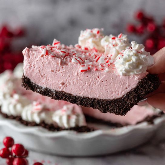 A slice of pink creamy pie with a dark brown crumbly crust is being lifted from a white pie dish. The pie has a thick pink filling sprinkled with crushed red and white peppermint pieces on top. Around the edges of the pie, there are dollops of white whipped cream, also topped with small pieces of peppermint. The background has bright red berries and a white marbled surface, adding a festive feel. Photo taken with an iphone --ar 4:5 --v 7