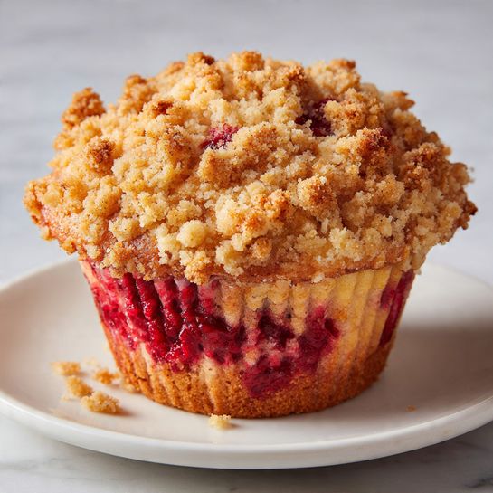 A single muffin sits centered on a white plate, placed on a white marbled surface. The muffin has two main layers: the bottom layer is a rich deep red color with a soft, moist texture, while the top layer is a crumbly light golden topping that looks crunchy and slightly uneven, with some crumbs falling onto the plate. The muffin is slightly rounded on top with the crumb topping spreading unevenly across it. Photo taken with an iphone --ar 4:5 --v 7