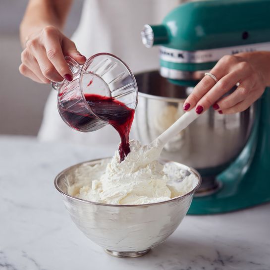 Velvet Blackberry Cake with Cream Cheese Frosting and Edible Flower Garnish Recipe 4 A woman’s hand holds a clear glass bowl pouring dark red thick liquid into a large silver mixing bowl filled with smooth, fluffy white cream. Another woman’s hand uses a white spatula to guide the dark red liquid into the cream. The mixing bowl is attached to a dark green mixer, all set on a white marbled surface. The scene focuses closely on the action of pouring and mixing. Photo taken with an iphone --ar 4:5 --v 7