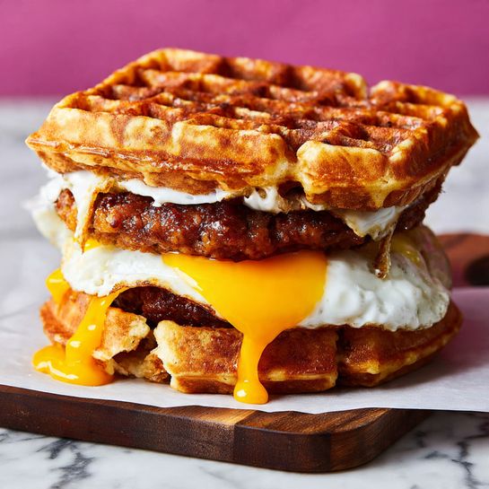 A sandwich with three main layers stacked between two waffle pieces that look golden brown and crispy. The bottom waffle is thick with a rough texture. Above it sits a dark brown fried patty, juicy and seasoned, topped with melted bright orange cheese that drips down the sides. On top of the cheese is a fried egg with a white set surface and bright yellow yolk spilling down the sandwich edges. The sandwich sits on white parchment paper over a dark wooden board, all placed on a white marbled surface with a bright, soft background. Photo taken with an iphone --ar 4:5 --v 7