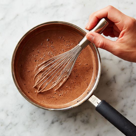 A close-up view of a stainless steel pan with a black handle placed on a white marbled surface, filled with a smooth, light brown chocolate mixture. A woman's hand is holding a metal whisk inside the pan, gently stirring the mixture. The light reflects softly on the surface of the chocolate mixture, showing its creamy texture. Photo taken with an iphone --ar 4:5 --v 7