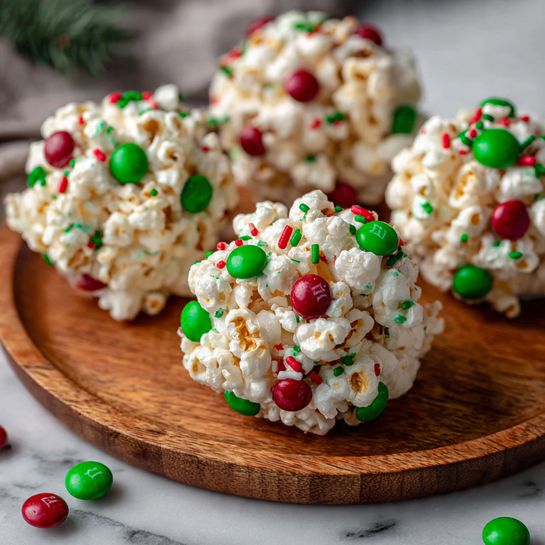 The image shows round popcorn balls with white popcorn as the main layer, tightly packed together to form a ball shape. Mixed in the popcorn are shiny red and green candy-coated chocolates scattered evenly throughout, along with small green and red sprinkles adding texture and color. The popcorn balls are arranged on a wooden plate resting on a white marbled surface. The popcorn looks light and slightly glossy, with the bright red and green candies standing out clearly against the pale popcorn. Photo taken with an iphone --ar 4:5 --v 7