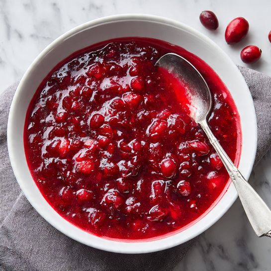 A white bowl filled with a thick, chunky red sauce made from crushed cranberries. The sauce has a shiny, slightly wet texture with visible large and small pieces of fruit. A silver spoon is dipped into the sauce on the right side of the bowl. The bowl is placed on a gray cloth with a few whole cranberries scattered around. The background shows a white marbled texture. Photo taken with an iphone --ar 4:5 --v 7