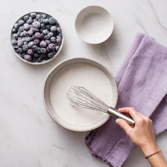 A round bowl filled with a light mixture being stirred by a woman's hand holding a silver whisk, the mixture is white with a slightly frothy texture on top. Above to the left, there is a round white plate filled with frozen blueberries covered in frost, showing dark purple and light blue shades. Above to the right of the bowl, there is a small empty white bowl. A light purple cloth is gently folded and placed to the right side of the image. All items are set on a white marbled surface. Photo taken with an iphone --ar 4:5 --v 7