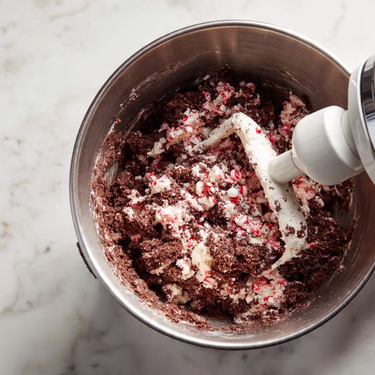 A close-up view from above shows a large silver mixing bowl filled with rough, dark brown cookie dough with small bits of red and white candy pieces mixed in. A white paddle attachment from a stand mixer is partly buried in the dough, coated with patches of the thick mixture. The bowl rests on a white marbled surface. The scene clearly shows the texture of the chunky dough and the contrast between the white paddle and the dark dough inside the shiny metal bowl photo taken with an iphone --ar 4:5 --v 7