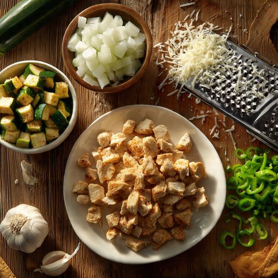 The image shows a wooden table with five main groups of food ingredients. At the bottom, there is a white plate filled with chopped cooked chicken pieces that have a light brown crust. Above the plate, to the left, there is a small bowl with roasted green zucchini cubes that have dark char marks. To the right of the zucchini, there is a pile of finely chopped white onions. Next to the onions and slightly above, there is a metal cheese grater with a large heap of grated white cheese beside it. On the right side of the image, there is a small pile of chopped green peppers. In the center lies a small garlic bulb with two peeled cloves sitting nearby. All items rest on a wooden surface. photo taken with an iphone --ar 4:5 --v 7