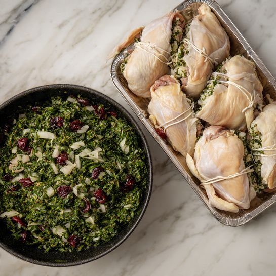 The first layer shows a round black pan filled with finely chopped green leafy herbs mixed with small white onion pieces and scattered dark red cranberries, all lightly cooked, placed on a white marbled surface. The second layer displays raw chicken pieces, tied with white string, arranged together on a silver tray, some showing green herb stuffing and a few cranberries peeking through the folds of the chicken skin, also placed on a white marbled surface. photo taken with an iphone --ar 4:5 --v 7
