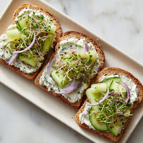 Three slices of toasted whole wheat bread are arranged side by side on a white rectangular plate, resting on a white marbled surface. Each toast slice is spread with a thick, creamy white mixture that has visible green herb pieces mixed in. On top of the spread are small, irregular slices of light green cucumber, thin curved strips of purple-red onion, and a generous handful of green sprouts, which add texture and a fresh look. The toppings look fresh and vibrant, with a slight shine from moisture. The photo is taken with an iphone --ar 4:5 --v 7