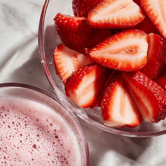 A close-up view shows a clear glass bowl filled with bright red, sliced strawberries, held above another clear glass bowl containing a light pink, frothy liquid. The strawberry slices show their red outer skin with tiny yellow seeds and pale flesh inside. The pink liquid below looks smooth and airy with small bubbles on the surface. The background is a white marbled texture. photo taken with an iphone --ar 4:5 --v 7