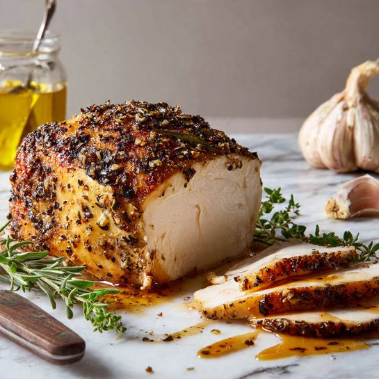 A large piece of cooked chicken with a brown, crispy skin speckled with black pepper and herbs sits in the center on a white marbled surface, showing a thick layer of white meat inside with green herbs between the skin and the meat. To the right is a smaller piece of sliced chicken with a similar brown crust and white inside. Near the bottom left, there is a browned garlic bulb with a few garlic cloves scattered around. Fresh green herb leaves lie close to the chicken. Behind the chicken is a glass jar with a golden liquid and a spoon inside. A large knife with a brown handle rests beside the chicken. In the far background, a separate chicken leg piece is partially visible. Photo taken with an iphone --ar 4:5 --v 7