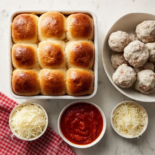A white square dish holds fifteen soft, golden-brown dinner rolls arranged in a 3 by 5 grid, each roll showing slight wrinkles on top and a fluffy texture. To the right, a white bowl contains round, pale gray meatballs coated lightly in flour with a rough texture. Below that, a small white bowl is filled with smooth, bright red marinara sauce topped with a subtle swirl pattern. In the bottom-left corner, a small white bowl holds a mound of shredded white mozzarella cheese with a slightly stringy texture. A red and white checkered cloth peeks in from the bottom left of the image, all placed on a white marbled textured surface. photo taken with an iphone --ar 4:5 --v 7