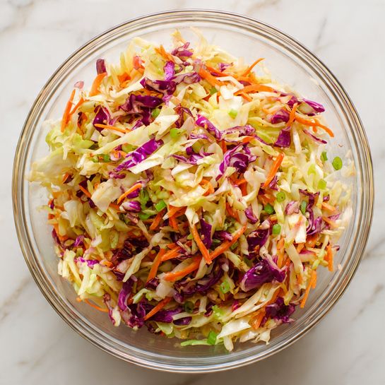 A clear glass bowl filled with a colorful mixed salad sits on a white marbled surface. The salad has many layers with shredded pale green cabbage, bright purple cabbage pieces, thin orange carrot strips, and small green bits that look like scallions or herbs, all mixed together in a light and fresh texture. The colors are vibrant and give a fresh, crunchy look. Photo taken with an iphone --ar 4:5 --v 7
