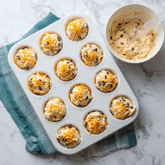 A white muffin tray with twelve round cavities, each filled with a mixture that has a creamy light beige base with small dark brown bits, scattered yellow shredded cheese, and small white chunks. The texture looks soft and wet, suggesting a batter before baking. The tray sits on a white marbled surface with a teal and white striped cloth partially underneath. In the upper right corner, a white bowl holds more of the same mixture, with a transparent plastic spoon inside. Photo taken with an iphone --ar 4:5 --v 7