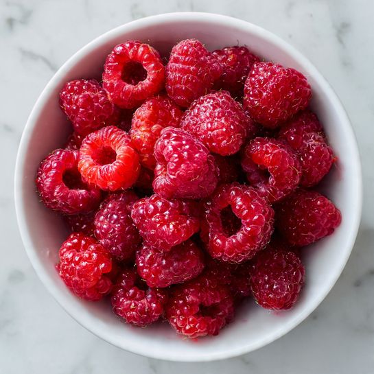 A white bowl filled with many fresh raspberries, some whole and some cut in half, showing their bright red color and small seed texture. The raspberries look juicy with some juice stains on the sides of the bowl. The bowl is placed on a white marbled surface. photo taken with an iphone --ar 4:5 --v 7