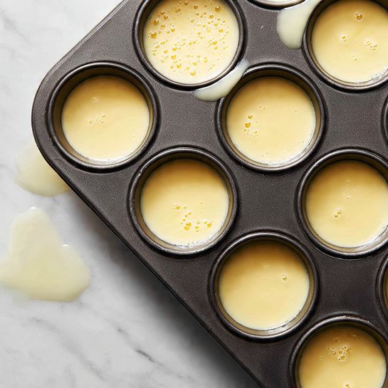 A close-up of a dark metal muffin tray with nine round cups filled with a smooth light yellow batter. Each cup is partially filled with the batter showing a creamy texture with small bubbles on the surface. The tray sits on a white marbled surface, and a bit of batter is spilled on the edges of a few cups, giving a natural messy look. The lighting is soft, highlighting the batter's glossy texture. photo taken with an iphone --ar 4:5 --v 7