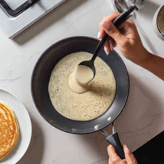 The image shows pancake batter being poured over three round dark chocolate pieces placed in the center of a black non-stick frying pan on a gas stove. The pancake batter layer is thick, light beige with small bubbles, covering the chocolate discs partially as it spreads evenly. A black spoon held by woman's hand is controlling the flow of the batter from above. To the left, a white plate with a cooked pancake sits on a white marbled surface. The background focuses on the pan and stove area, giving a close-up view of the cooking process photo taken with an iphone --ar 4:5 --v 7