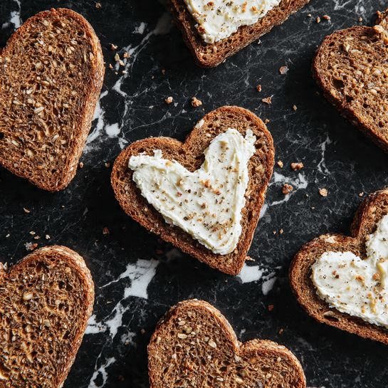 Several heart-shaped slices of brown bread with a creamy white spread on top are placed on a dark surface. The bread has a rough texture with visible grains, while the spread looks soft and slightly uneven. The slices are scattered casually, showing some crumbs around them. The background is a white marbled texture. Photo taken with an iphone --ar 4:5 --v 7