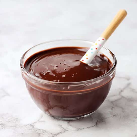A clear glass bowl filled with smooth, thick, dark brown chocolate sauce. The sauce has a shiny surface with small air bubbles. A white spatula with small colorful dots rests inside the bowl, covered halfway in the chocolate, with the tip dipped in the sauce. The bowl is placed on a white marbled surface for a clean and bright look. photo taken with an iphone --ar 4:5 --v 7