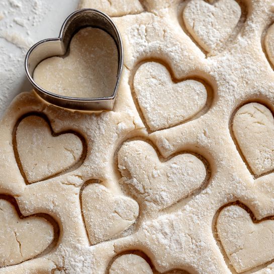 A close-up view of dough rolled flat in a light tan color with fine flour dusted on top, pressed with a metal heart-shaped cookie cutter partially lifted on the top left. The dough sheet shows many heart shapes, tightly packed in a neat pattern with smooth edges, ready to be separated and baked. The background is a white marbled texture. Photo taken with an iphone --ar 4:5 --v 7