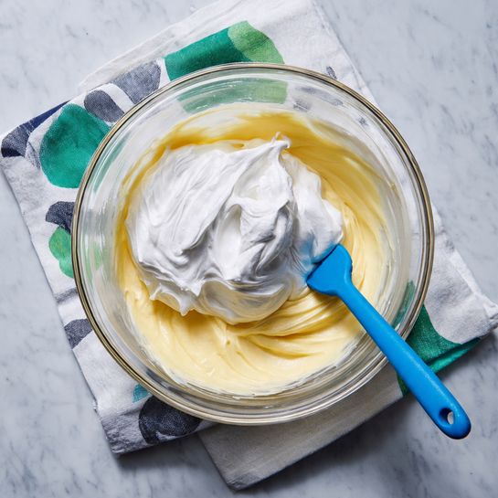 A clear glass bowl is shown with two layers inside: the bottom layer is a smooth, pale yellow creamy mixture with swirled texture, and on top is a dollop of thick, white whipped cream with soft peaks. A blue silicone spatula is partially placed in the bowl, resting on the whipped cream. The bowl is placed on a folded white cloth with green and black abstract patterns, all set on a white marbled surface. Photo taken with an iphone --ar 4:5 --v 7