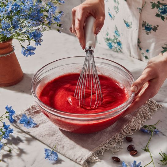 A clear glass bowl filled with smooth, thick red batter is being stirred with a silver whisk that has a white handle. The bowl sits on a cloth over a white marbled surface, scattered with small blue flowers and coffee beans around it. One woman's hand holds the bowl steady on the right side while another woman's hand grips the whisk on the left. The background features a white apron with delicate blue floral prints, and a terra-cotta vase filled with small blue flowers is visible to the left. The light is soft, casting slight shadows across the scene. photo taken with an iphone --ar 4:5 --v 7