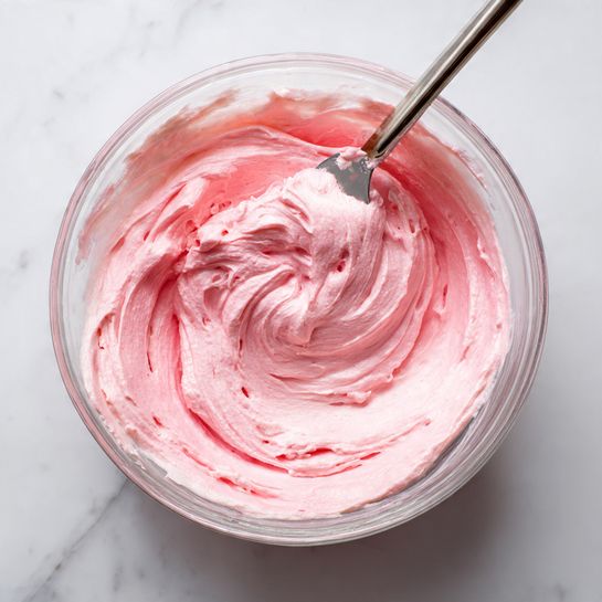 A clear glass bowl filled with smooth, thick pink frosting swirled inside, showing shades of light and medium pink spread evenly along the sides. A metal spatula with some frosting on it rests inside the bowl, angled slightly upward. The bowl sits on a white marbled surface, emphasizing the creamy texture and vibrant pink color of the frosting. Photo taken with an iphone --ar 4:5 --v 7