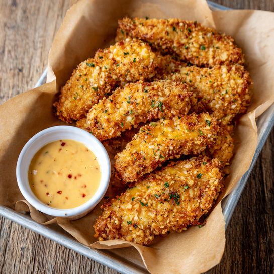 The image shows a tray lined with light brown parchment paper holding seven pieces of golden-brown crispy fried chicken tenders. The chicken tenders have a crunchy texture with an uneven coating, sprinkled lightly with small green herb bits. In the bottom left corner of the tray, there is a small white cup filled with creamy, light orange dipping sauce with some small red specks. The tray sits on a wooden surface. photo taken with an iphone --ar 4:5 --v 7