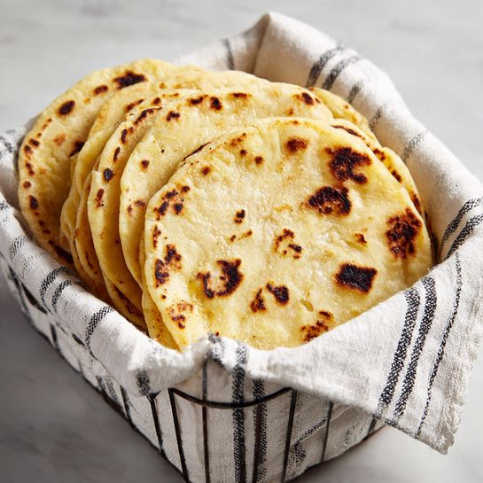 A white cloth with black stripes lines a container filled with a stack of round, light yellow corn tortillas showing uneven edges and some brown char marks on top, with slight texture from the cooking spots on each tortilla; the container sits on a white marbled surface. photo taken with an iphone --ar 4:5 --v 7