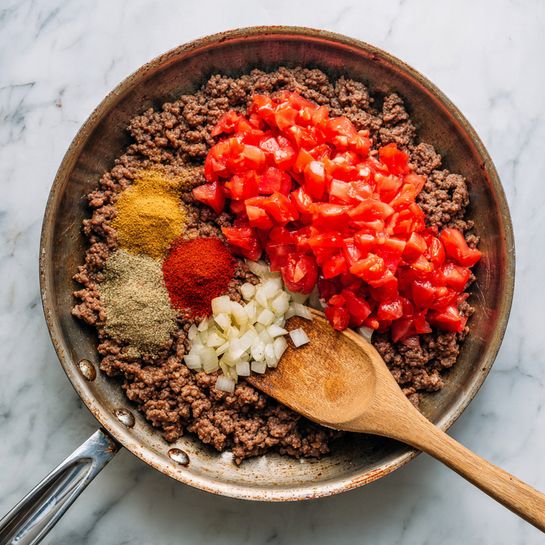 A round metal pan with browned ground meat and small pieces of cooked onion spread evenly around the bottom forms the base layer. On top of the meat, there are two piles of vibrant red chopped tomatoes placed side by side towards one side of the pan. Near the tomatoes, there are three small mounds of spices in different colors: one deep red, one light brown, and one yellowish-brown. A wooden spatula rests partly on the tomatoes and spices, with some diced onion on it. The pan handle is visible at the bottom, and the whole scene sits on a surface with a white marbled texture photo taken with an iphone --ar 4:5 --v 7