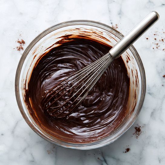 A clear glass bowl filled with dark brown, smooth chocolate batter, showing thick and shiny texture, with a metal whisk resting inside. The bowl sits on a white marbled surface that has small splashes and smudges of cocoa powder scattered around. The batter clings slightly to the edges of the bowl, with some streaks made by the whisk. Photo taken with an iphone --ar 4:5 --v 7