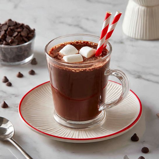A clear glass cup filled with dark brown hot chocolate topped with two large white marshmallows floating on the surface. A red and white striped straw is placed inside the cup, leaning slightly to the left. The cup sits on a round white plate with a ridged edge, all set against a white marbled surface. There are scattered chocolate chips around the plate and a silver spoon lies in the foreground. In the background, there is a blurred white ceramic figure and a small container filled with more chocolate chips. Photo taken with an iphone --ar 4:5 --v 7