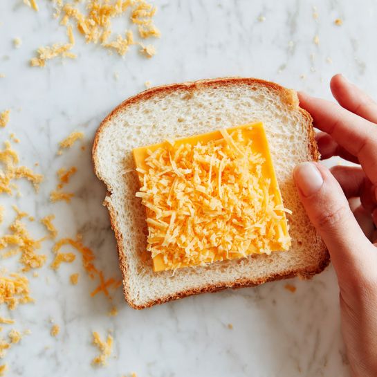 A piece of soft white bread is laid flat with its crust removed, showing a single square slice of orange cheddar cheese placed in the center. The bread is partially rolled up from the right side by a woman's hand, with fingers gently curling the bread inward. The background is a white marbled surface with some small bread crumbs scattered around. photo taken with an iphone --ar 4:5 --v 7