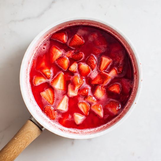 A top view of a white saucepan with a wooden handle containing a mixture of chopped strawberries simmering in a thick red liquid. The strawberries are cut into uneven pieces showing bright red colors with some foam bubbles on the surface of the juice. The edges of the pan show red splashes from the cooking process. The saucepan is placed on a white marbled surface. photo taken with an iphone --ar 4:5 --v 7