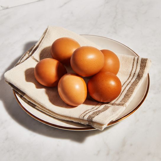 Eight shiny brown eggs are placed in two rows, four on top and four below, on top of a beige cloth with two thin brown stripes near the edge. The cloth sits inside a white plate with a bronze rim. The whole scene rests on a white marbled surface. The eggs have smooth textures and a warm orange-brown color that catches the light in soft reflections. photo taken with an iphone --ar 4:5 --v 7