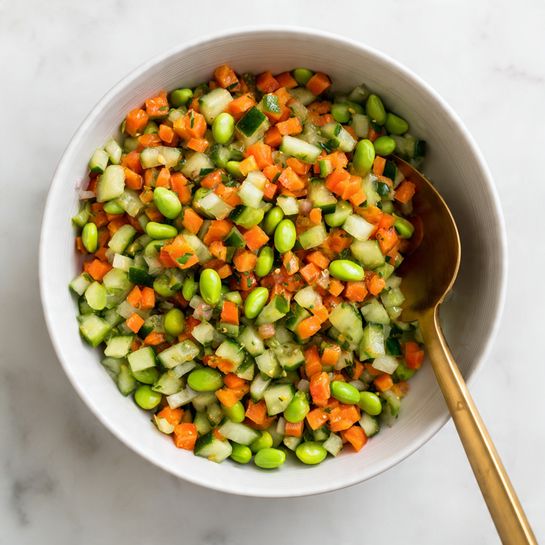 Roll Baked Sushi Cups Recipe 4 A white bowl filled with a colorful mix of chopped vegetables, including bright green edamame beans, diced light green cucumbers, and small pieces of orange carrots. The vegetables are cut into small, bite-sized chunks and spread evenly throughout the bowl. A golden spoon is placed inside the bowl, with its handle resting on the edge, partially covered by the vegetables. The bowl sits on a white marbled surface, adding a clean and fresh look to the scene. photo taken with an iphone --ar 4:5 --v 7
