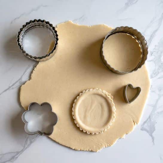 A layer of light beige dough rolled flat on a white marbled surface, with two round scalloped dough shapes cut out and placed nearby. There are two metal cookie cutters, one placed on the dough and the other beside it; the cutter on the dough is round with scalloped edges, and the other has a small heart shape inside a larger scalloped circle shape. There is a partial cut where a round scalloped shape was taken from the dough, showing the light beige texture against the smooth white marbled surface photo taken with an iphone --ar 4:5 --v 7