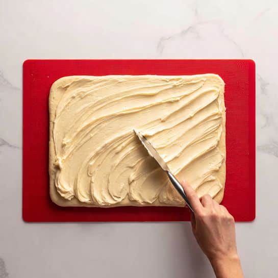 A rectangular piece of dough, light beige in color and soft in texture, lies flat on a red silicone baking mat placed on a white marbled surface. On the left side of the dough, a thick, creamy, pale yellow layer of spread is being evenly smoothed with a metal spatula held by a woman's hand. The spread covers about one-third of the dough’s surface and shows textured, smooth waves from the spreading motion. Photo taken with an iphone --ar 4:5 --v 7