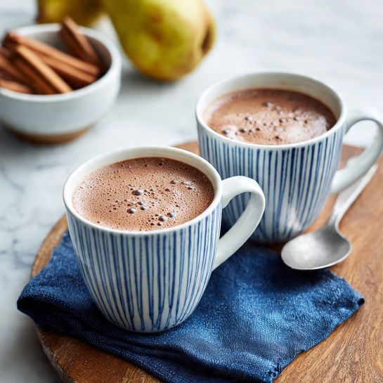 A white cup with blue stripes holds smooth, light brown hot chocolate, with a steady, thin stream pouring into it, creating small bubbles on the surface. The cup sits on a folded blue napkin on a wooden table with a silver spoon resting beside it. In the background, a white bowl filled with green pears is softly out of focus, alongside another bowl and cup. The scene is set on a white marbled surface. photo taken with an iphone --ar 4:5 --v 7