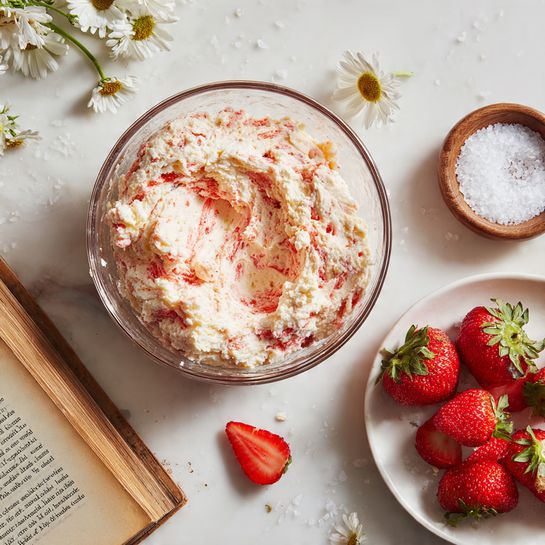Strawberry Cheesecake Cookies Recipe 4 A clear glass bowl filled with a dough-like mixture that is cream-colored with streaks of bright red swirled throughout, showing a textured and slightly crumbly look. Around the bowl, there is a white marbled surface with some scattered crumbs. To the bottom right, a white plate holds several fresh, glossy red strawberries with green leaves. Near the plate, two strawberry halves lie on the white marbled surface. To the left, there is an open book with aged yellow pages and a small round wooden bowl filled with white granulated sugar resting on the pages. Small white daisy flowers add a delicate touch near the strawberries and book. Photo taken with an iphone --ar 4:5 --v 7